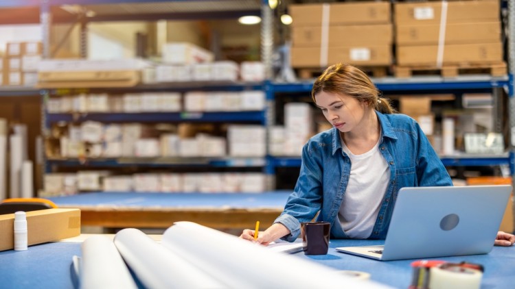 Woman Using Laptop In Warehouse For Management